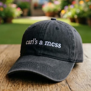 May include: Black baseball cap with a curved brim. The cap features the embroidered text "carl's a mess" in white. The hat is made of a soft, worn material and sits on a wooden surface.