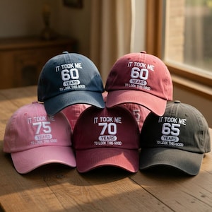 May include: A collection of six baseball caps in various colors, including navy, red, pink, burgundy, and charcoal. Each cap features the embroidered phrase "IT TOOK ME [number] YEARS TO LOOK THIS GOOD."