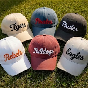 May include: Six baseball caps in various colors, including tan, navy, maroon, black, and white. Each cap features embroidered text: "Tigers," "Pirates," "Rebels," "Bulldogs," "Eagles," and "Pirates."
