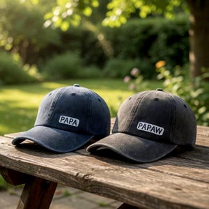 May include: Two baseball caps with curved brims rest on a wooden surface. One is navy blue with the word "PAPA" in white, and the other is charcoal gray with "PAPAW" in white. The caps are in a natural outdoor setting.