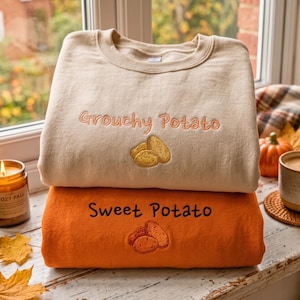 May include: A beige crewneck sweatshirt with the embroidered text "Grouchy Potato" above a yellow potato graphic, stacked on an orange crewneck sweatshirt with the embroidered text "Sweet Potato" above an orange potato graphic.