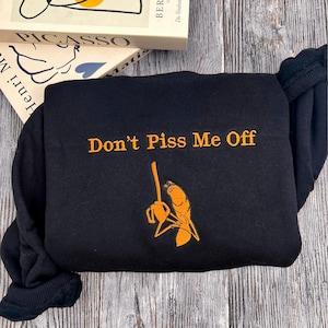 May include: Black sweatshirt with the embroidered phrase "Don't Piss Me Off" in orange. Below the text is an orange graphic of a person holding a mop. The sweatshirt is folded on a wooden surface.