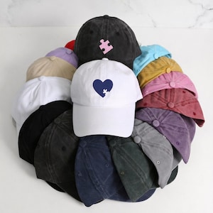 May include: A collection of baseball caps in various colors, including white, black, and shades of blue, pink, and purple. The caps feature a curved brim and adjustable strap. Some caps have embroidered designs, such as a heart with a puzzle piece.