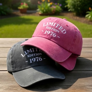 May include: A pink baseball cap and a dark gray baseball cap, both featuring white embroidered text that reads LIMITED EDITION - 1976 - on the front panels. Each cap has a curved brim and a structured crown with a button on top.