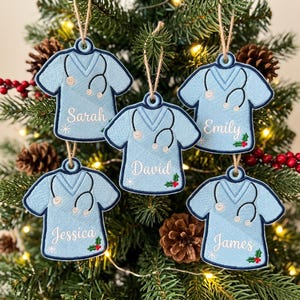 May include: Five light blue scrub top ornaments with dark blue trim, each featuring an embroidered black stethoscope, a small snowflake, and a sprig of holly. The names Sarah, David, Emily, Jessica, and James are stitched in white script.