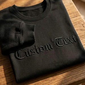 May include: Black sweatshirt with the embroidered text "Custom Text" in a gothic font. The sweatshirt is displayed on a wooden surface, showcasing the crew neck and ribbed cuffs.