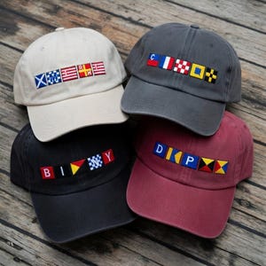 May include: A collection of five baseball caps in various colors, including beige, gray, black, and burgundy. Each cap features embroidered nautical signal flags spelling out different letter combinations. The caps are arranged on a wooden surface.