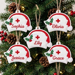 May include: Five white and red nurse cap ornaments hanging from twine. Each cap features a red cross, a green holly leaf with red berries, and a personalized name in red script: Sophia, Olivia, Lily, Jessica, and Grace. The ornaments are shaped like traditional nurse hats.
