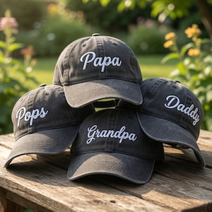 May include: Four dark gray baseball caps with white embroidered text: "Papa", "Pops", "Daddy", and "Grandpa". The caps are arranged on a wooden surface, with a blurred green background.