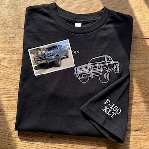 May include: Black t-shirt featuring a photograph of a silver pickup truck and a white embroidered outline of a truck. The sleeve has the text "F-150 XLT" in white.