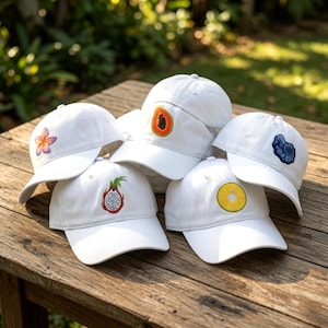 May include: Five white baseball caps arranged together, each featuring a unique embroidered design on the front panel: a pink and yellow plumeria flower, an orange papaya slice, a blue seashell, a dragon fruit, and a yellow pineapple slice.