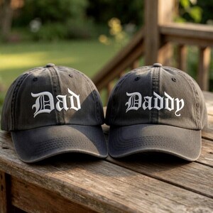 May include: Two charcoal gray baseball caps with curved visors. One cap has the word "Dad" embroidered in white, the other reads "Daddy". The caps are displayed on a wooden surface, suggesting a casual, outdoor setting.