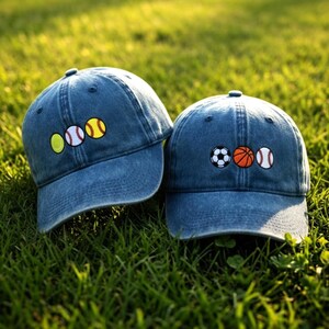 May include: Two denim baseball caps embroidered with sports ball designs. One cap features a tennis ball, baseball, and a softball. The other cap displays a soccer ball, basketball, and baseball. The caps are on a green grassy surface.