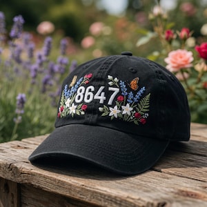 May include: Black baseball cap with embroidered floral design and the number 8647 in white. The embroidery includes butterflies, white flowers, red roses, and blue sprigs. The cap is sitting on a wooden surface.