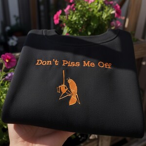 May include: Black sweatshirt with the embroidered phrase "Don't Piss Me Off" in orange thread. Below the text is an orange graphic of a bird holding a fishing rod. The sweatshirt is a casual garment.
