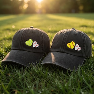 May include: Two black baseball caps with heart-shaped patches. One cap has a tennis ball heart and a baseball heart. The other cap has a softball heart and a baseball heart. The caps are on green grass.