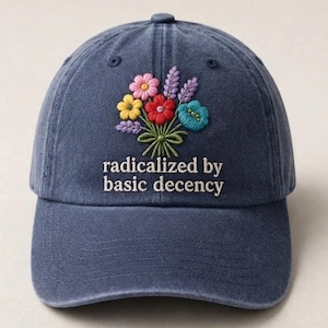 May include: A navy blue baseball cap featuring an embroidered bouquet of colorful flowers in pink, yellow, red, purple, and teal. Below the floral design, the text "radicalized by basic decency" is embroidered in white, sans-serif lettering.