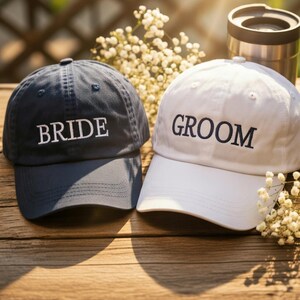 May include: Two baseball caps, one navy blue and one white, sit side-by-side on a wooden surface. The navy cap has the word "BRIDE" embroidered in white, while the white cap says "GROOM" in black. Small white flowers are scattered around the caps.
