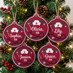 May include: Five round red Christmas ornaments featuring a white nurse cap with a red cross, a black stethoscope, and the names Sophia, Olivia, Lily, Jessica, and Grace followed by RN. Each ornament has a green pine and berry graphic.