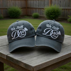 May include: Two gray baseball caps with the text "We Still D 40 years" embroidered in white. The caps are sitting on a wooden table outdoors. The caps have a curved brim and a top button.