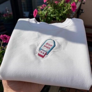 May include: White sweatshirt with an embroidered USB drive design. The USB drive has a blue outline, a red connector, and the words "JESUS SAVES" embroidered in blue and red.