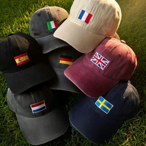 May include: A collection of baseball caps in various colors, each featuring a small embroidered flag. Flags include Spain, Italy, France, Germany, the United Kingdom, Sweden, and the Netherlands. The caps are stacked on green grass.