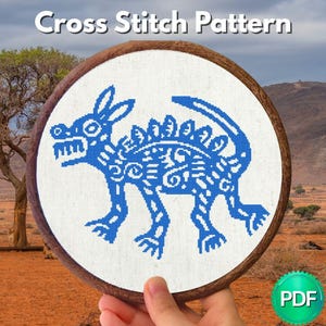 May include: A cross stitch pattern featuring a blue, stylized creature with a long tail and spiky back, embroidered on white fabric within a circular wooden hoop. The text "Cross Stitch Pattern" appears at the top, and a green "PDF" icon is visible.