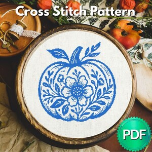 May include: A cross stitch pattern featuring a blue pumpkin design with floral details, displayed in a wooden embroidery hoop. The text "Cross Stitch Pattern" is at the top. A PDF icon is in the bottom right corner.