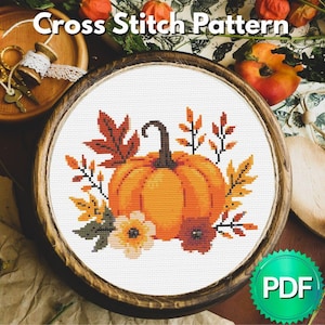 以下が含まれることがあります： 木製の刺繍枠に入った、オレンジ色のカボチャと秋の葉と花をあしらったクロスステッチのデザイン。「Cross Stitch Pattern」の文字が上部にあります。右下にはPDFアイコンがあります。