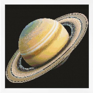 May include: A cross-stitch embroidery of the planet Saturn, featuring a spherical body in shades of yellow, tan, and pale blue, surrounded by prominent rings in beige and grey tones, all set against a solid black background within a white frame.