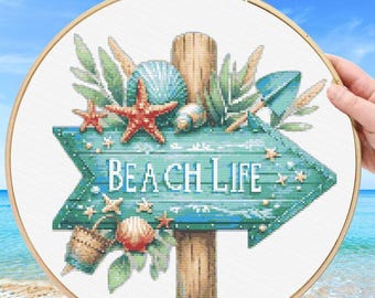 May include: A cross stitch pattern PDF featuring a wooden signpost shaped like an arrow pointing right with the text BEACH LIFE. The design includes a red starfish, various seashells, green palm leaves, and a woven basket on a white fabric background.