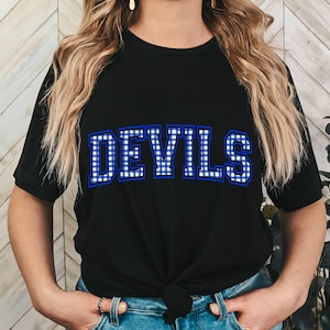 Puede incluir: Camiseta negra con la palabra "DEVILS" en letras grandes a cuadros azules y blancos en el pecho. La camiseta tiene mangas cortas y cuello redondo. La modelo lleva vaqueros azules.
