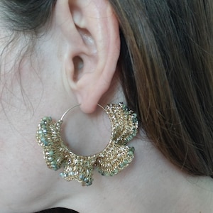 Puede incluir: Un pendiente de aro grande con una estructura de alambre dorado metálico adornado con un intrincado tejido de ganchillo con volantes. El diseño incorpora pequeñas cuentas brillantes de color verde salvia tejidas en el hilo dorado, creando un accesorio circular texturizado.
