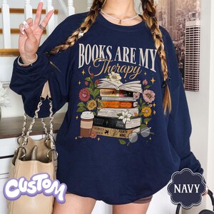 May include: Navy blue sweatshirt with the text "BOOKS ARE MY Therapy" above a stack of books, flowers, and a coffee cup. The design includes glasses and stars. The word "NAVY" is in a cloud-shaped tag.