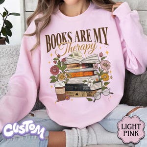 May include: Light pink sweatshirt with the text "BOOKS ARE MY Therapy" in brown script. The graphic features a stack of books, flowers, a coffee cup, and glasses. The bottom right corner has a "LIGHT PINK" label.