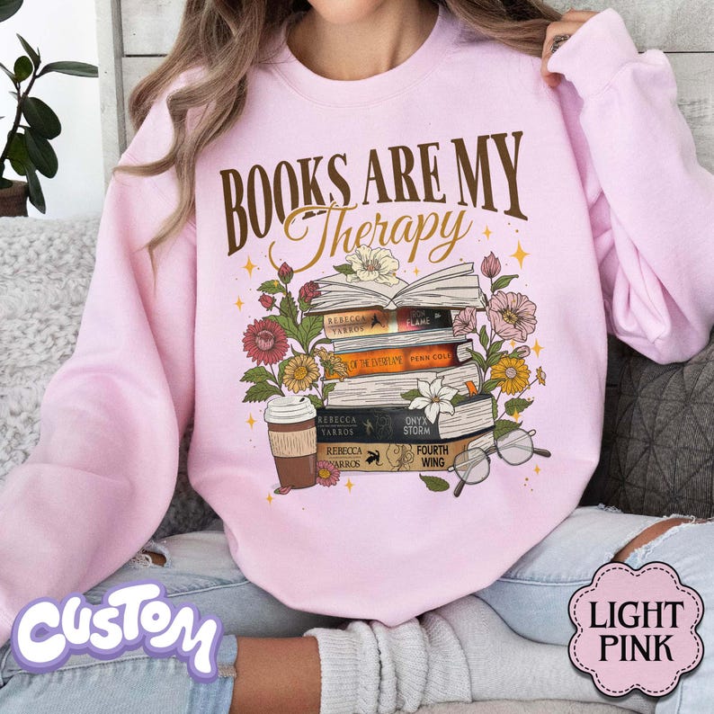 May include: Light pink sweatshirt with the text "BOOKS ARE MY Therapy" in brown script. The graphic features a stack of books, flowers, a coffee cup, and glasses. The bottom right corner has a "LIGHT PINK" label.