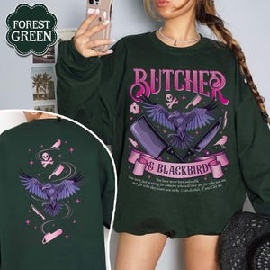 May include: Forest green sweatshirt with a graphic design featuring a raven, cleavers, and the text "BUTCHER & BLACKBIRD". The design includes a banner with text and additional illustrations. The back of the sweatshirt also has a similar design.