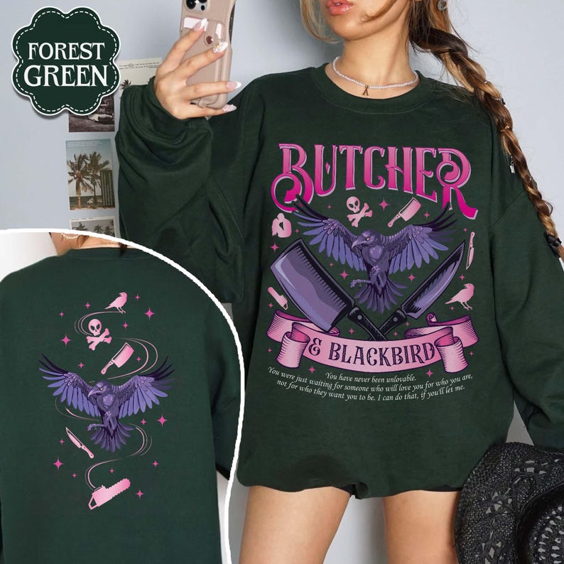 May include: Forest green sweatshirt with a graphic design featuring a raven, cleavers, and the text "BUTCHER & BLACKBIRD". The design includes a banner with text and additional illustrations. The back of the sweatshirt also has a similar design.