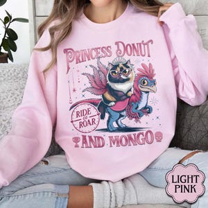 May include: Light pink sweatshirt with a graphic featuring a cat wearing a crown riding a dinosaur. The text reads "Princess Donut" above the image and "Ride or Roar" and "And Mongo" below.