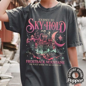May include: Dark gray t-shirt with a fantasy graphic. The design features a castle, dragon, and crescent moon in pink and green hues, with the text "JOURNEY TO SKY HOLD" and "FROSTBACK MOUNTAINS". The shirt has a vintage look.