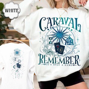 K&ouml;nnte beinhalten: Wei&szlig;es Sweatshirt mit einem teal- und blaugr&uuml;nen Grafikdesign. Das Design enth&auml;lt das Wort "CARAVAL" &uuml;ber dem Wort "REMEMBER" und den Satz "IT'S ONLY A GAME". Die R&uuml;ckseite des Sweatshirts hat ein &auml;hnliches Design.