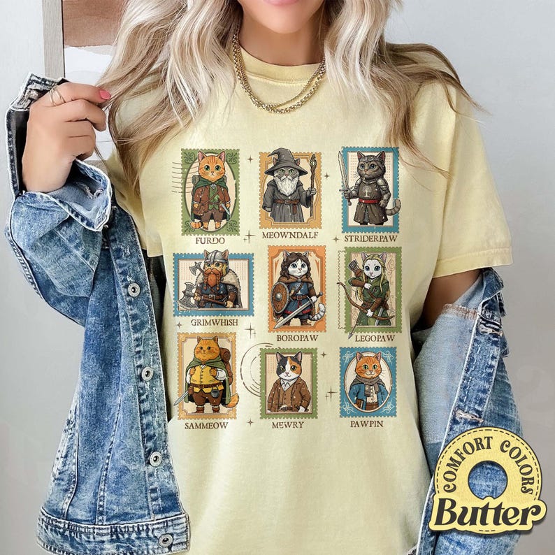 K&ouml;nnte beinhalten: Hellgelbes T-Shirt mit neun Katzenillustrationen in Briefmarkendesigns. Jede Katze ist als Figur mit Namen wie Furdo, Meowndalf und Striderpaw gekleidet. Das Shirt hat ein Comfort Colors Butter-Label.