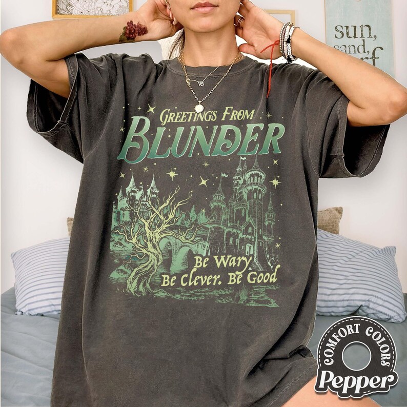 Puede incluir: Camiseta gris oscuro con un dise&ntilde;o desgastado. El dise&ntilde;o incluye el texto "Greetings From Blunder" sobre una ilustraci&oacute;n de un castillo. Debajo, el texto "Be Wary, Be Clever, Be Good". Marca Comfort Colors.