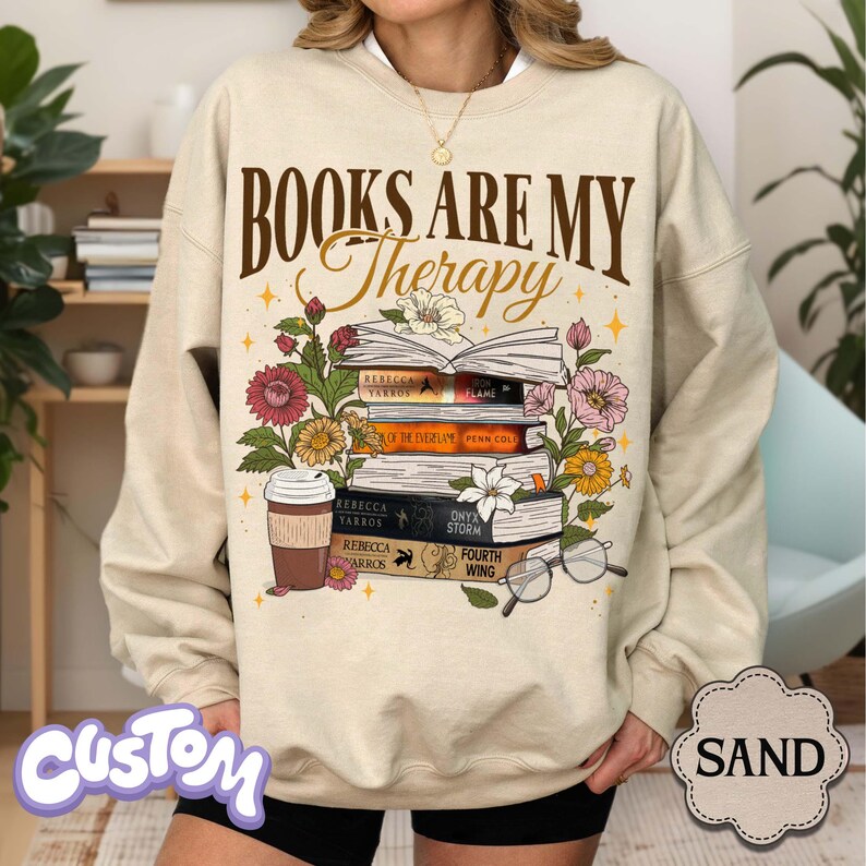 May include: Sand-colored sweatshirt with the text "BOOKS ARE MY Therapy" above a stack of books, flowers, and a coffee cup. The books have titles like "FLAME" and "ONYX STORM." The sweatshirt has a crew neck and long sleeves.