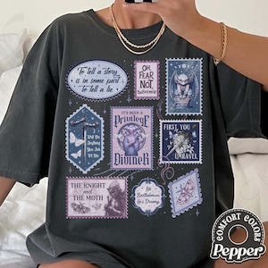 May include: Dark grey t-shirt with multiple postage stamp-style graphics. The stamps feature illustrations of a bat, butterflies, a sword, and text including "DivineR" and "The Knight and the Moth." The shirt has the "Comfort Colors" and "Pepper" logo.