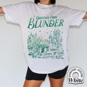 Puede incluir: Camiseta blanca con un gr&aacute;fico verde que dice "Greetings From Blunder" sobre una ilustraci&oacute;n de un castillo. El gr&aacute;fico tambi&eacute;n incluye las frases "Be Wary, Be Clever, Be Good". La camiseta tiene un logotipo redondo Comfort Colors White.