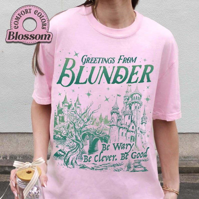 Puede incluir: Una camiseta rosa claro con el texto "Greetings From Blunder" en verde, junto con una ilustraci&oacute;n de un castillo. La camiseta tambi&eacute;n tiene las frases "Be Wary, Be Clever, Be Good" en verde. La camiseta tiene mangas cortas.