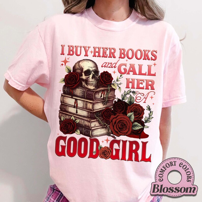 May include: Light pink t-shirt with a graphic design featuring a skull, roses, and stacked books. The text reads "I BUY HER BOOKS and GALL HER A GOOD GIRL" in red. The shirt has a Comfort Colors label.