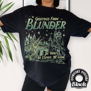 Puede incluir: Camiseta negra con un gr&aacute;fico verde y blanco que dice "Greetings From Blunder" sobre una ilustraci&oacute;n de un castillo. El texto "Be Wary, Be Clever, Be Good" est&aacute; debajo del castillo. La camiseta tiene una etiqueta Comfort Colors.