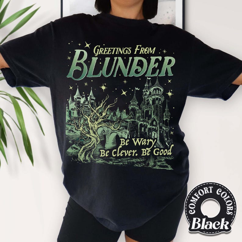 Puede incluir: Camiseta negra con un gr&aacute;fico verde y blanco que dice "Greetings From Blunder" sobre una ilustraci&oacute;n de un castillo. El texto "Be Wary, Be Clever, Be Good" est&aacute; debajo del castillo. La camiseta tiene una etiqueta Comfort Colors.